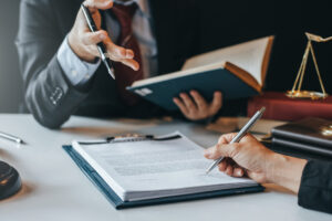 Two professionals sign a document at a desk, one writing while the other points with a pen beside a blue binder and legal books.