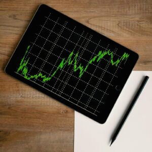 Tablet showing a bright green stock chart on a black screen, angled on a wooden desk next to a white sheet and black pen.