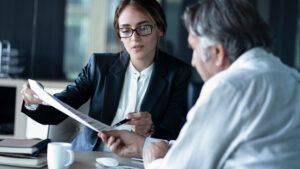 Businesswoman in a blazer and glasses reviews a document with a male colleague at a conference table.