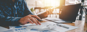 Hands typing on a calculator while reviewing financial charts at a wooden desk with a laptop nearby.