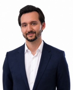 Professional headshot: man with dark hair and a short beard, wearing a navy suit and white shirt, smiling slightly against a white background.