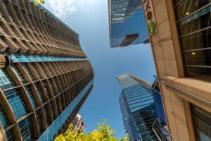 Low-angle view of tall glass and concrete skyscrapers forming a narrow urban canyon against a blue sky, with green trees at the bottom.