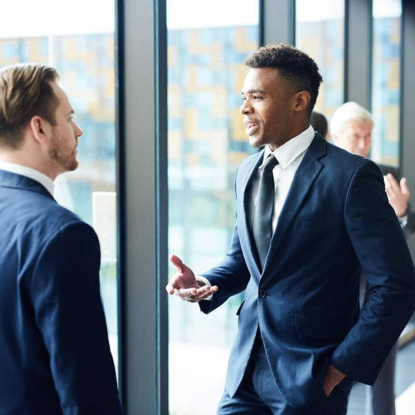 Man in a navy suit talks with a colleague beside large windows in a bright office hallway.