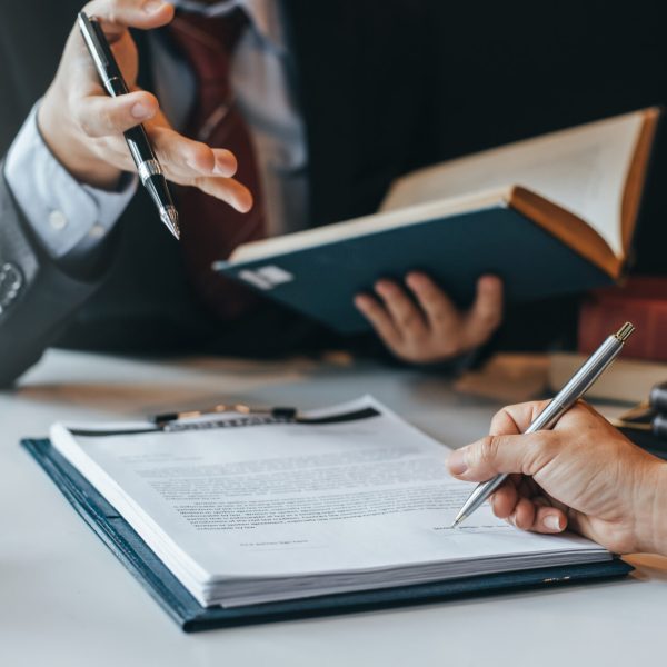 Two professionals sign a document at a desk, one writing while the other points with a pen beside a blue binder and legal books.